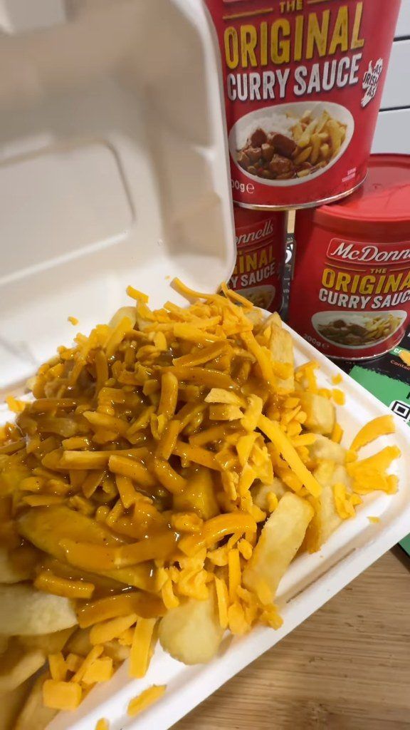 A box of chips loaded with grated cheese and curry sauce, with tins of McDonnells curry sauce behind